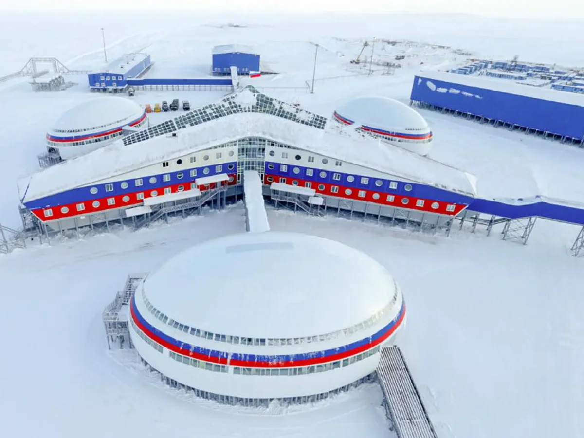 Vista aérea de la base militar rusa "Trébol Ártico" en la Isla Alexandra. El complejo principal tiene una forma geométrica de trébol con tres alas unidas a un núcleo central, pintado con franjas horizontales de color blanco, azul y rojo. La estructura está rodeada por un paisaje completamente cubierto de nieve blanca bajo un cielo nublado, con otros edificios industriales azules y pasarelas elevadas en el fondo.