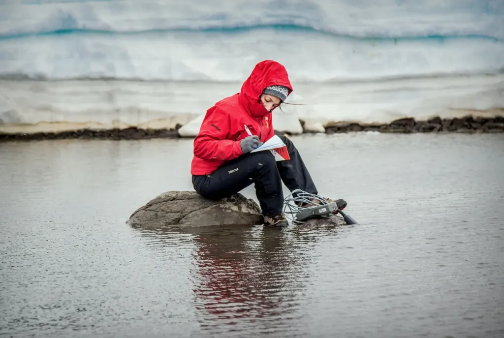 Investigadora antártica chilena toma notas de campo junto a un instrumento de muestreo, sentada sobre una roca frente a un glaciar.