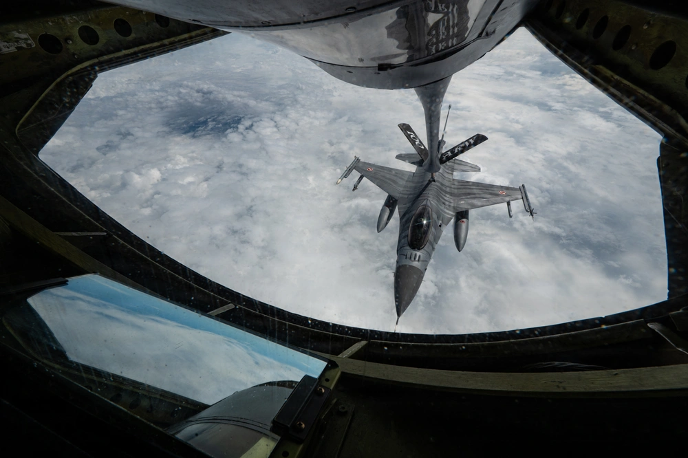 Un caza de combate recibe combustible en pleno vuelo desde un avión cisterna sobre una espesa capa de nubes, visto a través de la ventana del operador de reabastecimiento.