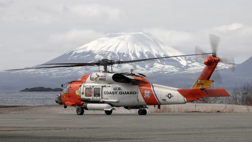 Helicóptero MH-60T Jayhawk de la Guardia Costera de Estados Unidos preparándose para despegar en una pista de Sitka, Alaska, con montañas nevadas en el fondo.
