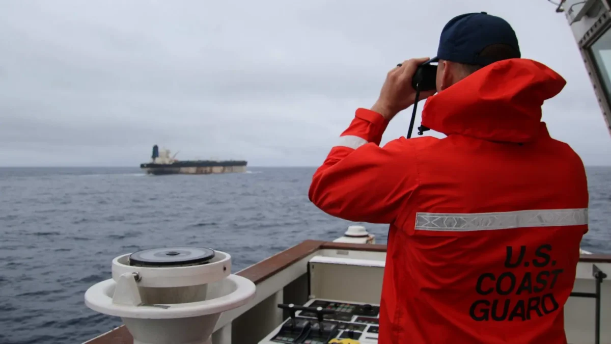 Un oficial de la Guardia Costera de Estados Unidos, vestido con chaqueta roja de alta visibilidad, observa a través de binoculares a un buque navegando en el horizonte.