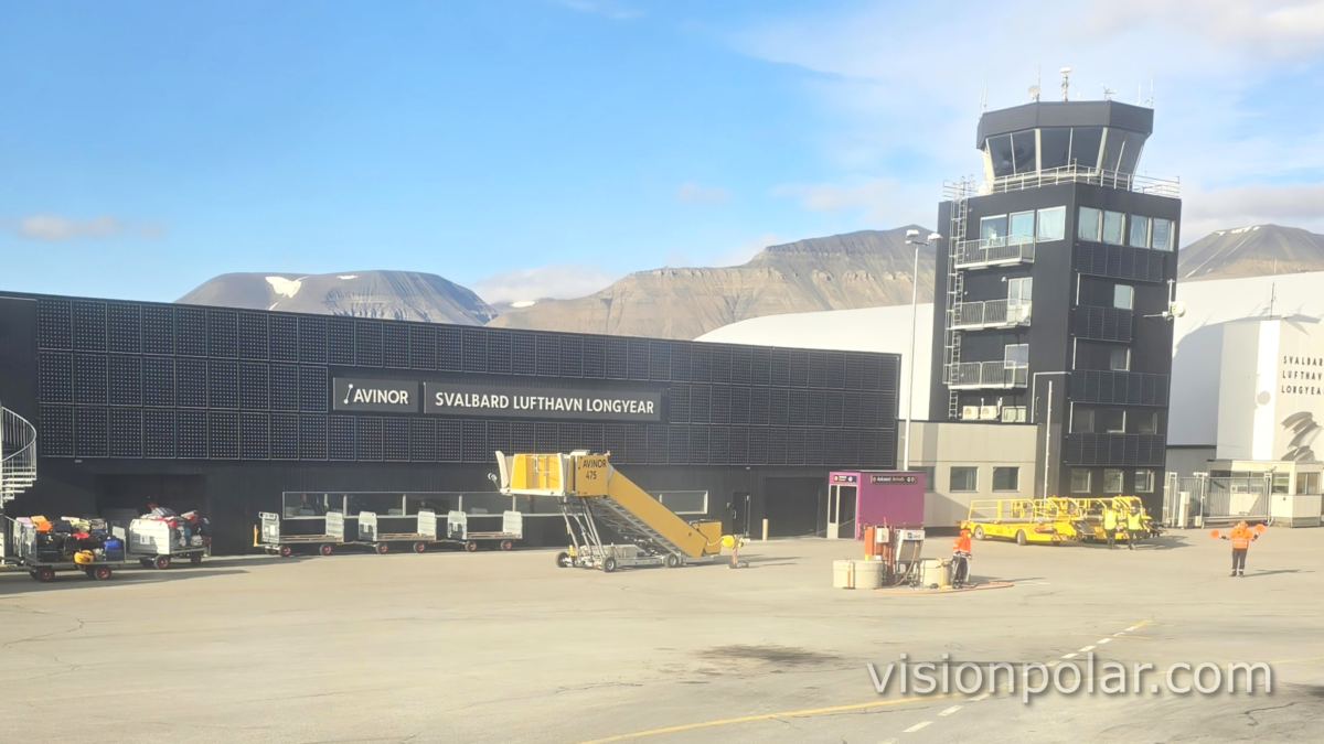 Terminal y torre de control del aeropuerto de Svalbard, en Longyearbyen, con el cartel de Avinor y montañas árticas al fondo bajo cielo despejado.