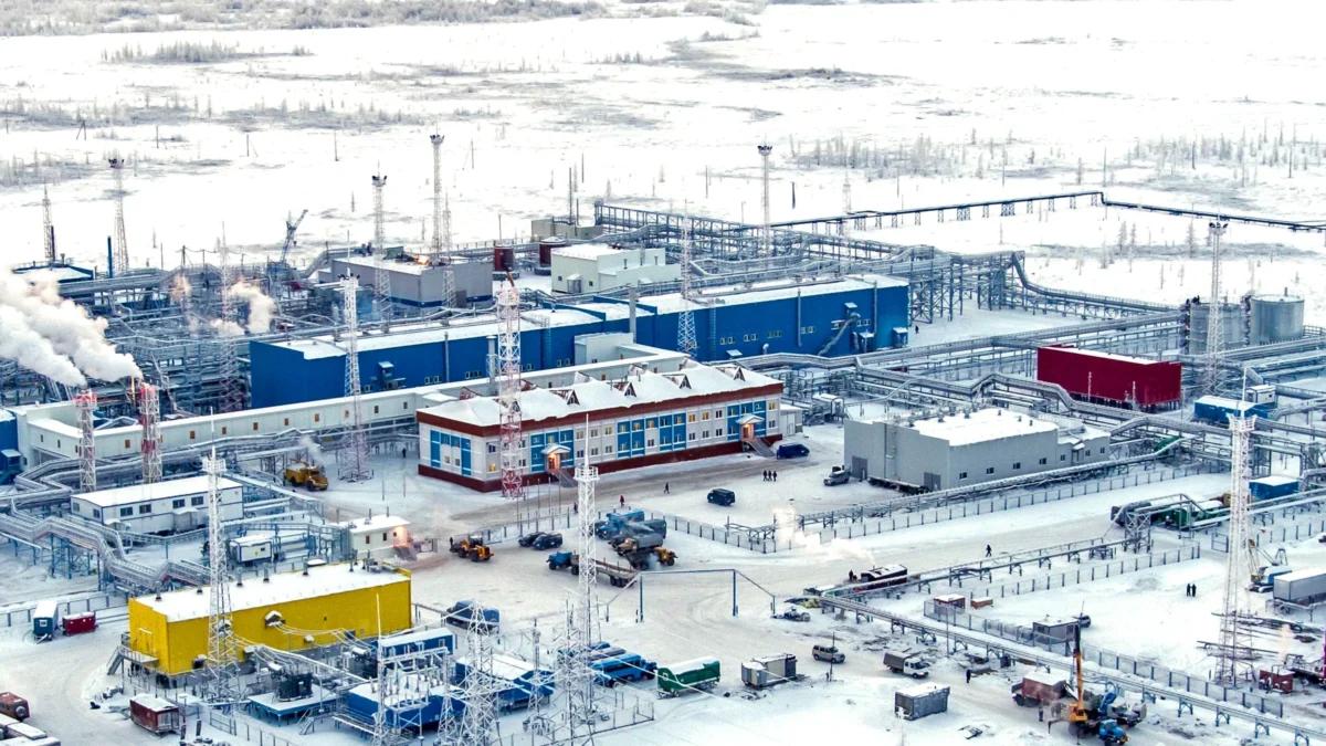 Vista aérea de una planta de procesamiento de gas rodeada de nieve en la tundra ártica, con edificios industriales azules, torres de iluminación y tuberías.