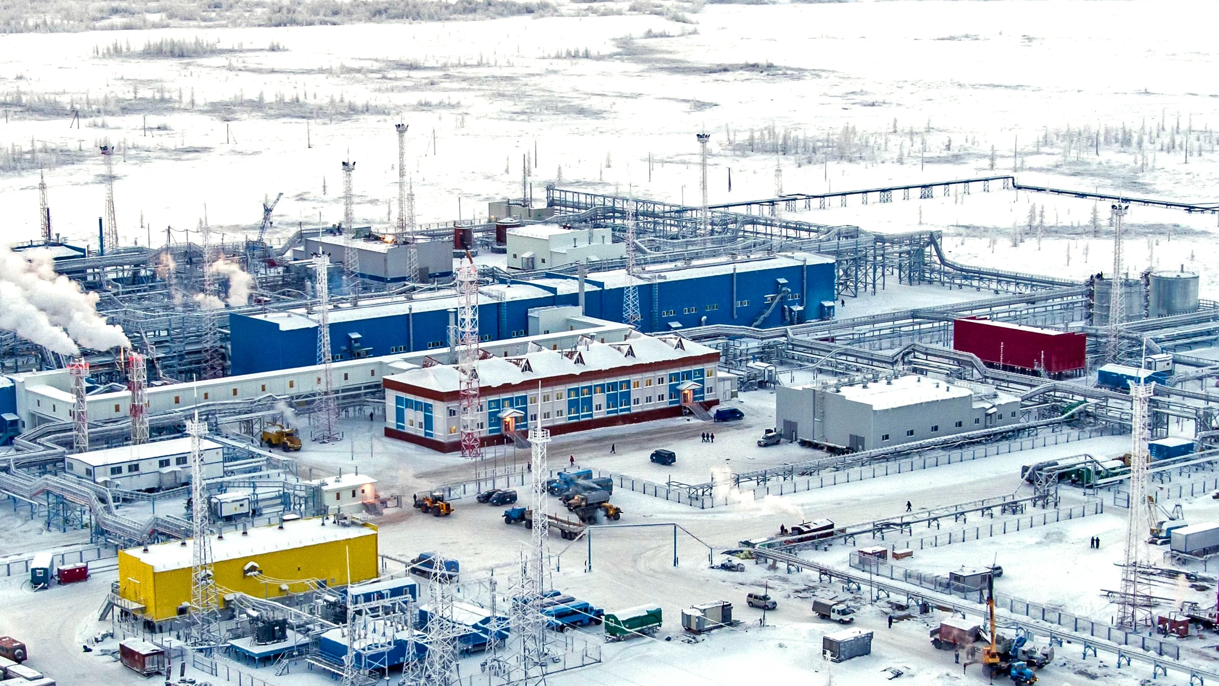 Vista aérea de una planta de procesamiento de gas rodeada de nieve en la tundra ártica, con edificios industriales azules, torres de iluminación y tuberías.