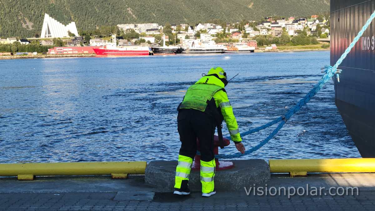Operario de amarre con ropa de alta visibilidad asegura un cabo azul en un noray del muelle de Tromsø; al fondo, cruzando el Tromsøysundet, la Catedral Ártica y las montañas de Tromsdalen.