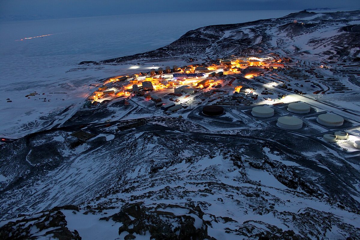 Vista aérea de la estación McMurdo al anochecer antártico, con las luces de los edificios iluminando el terreno volcánico de la isla Ross.